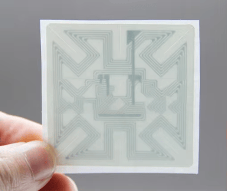 Deze afbeelding toont een vierkante RFID-inlay die met de hand wordt vastgehouden, waarbij het geleidende antennecircuit en de centrale chipstructuur duidelijk zichtbaar zijn.